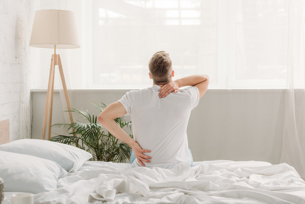 Вид сзади человека, сидящего на белом постельном белье и страдающего от боли в спине
 - Фото, изображение