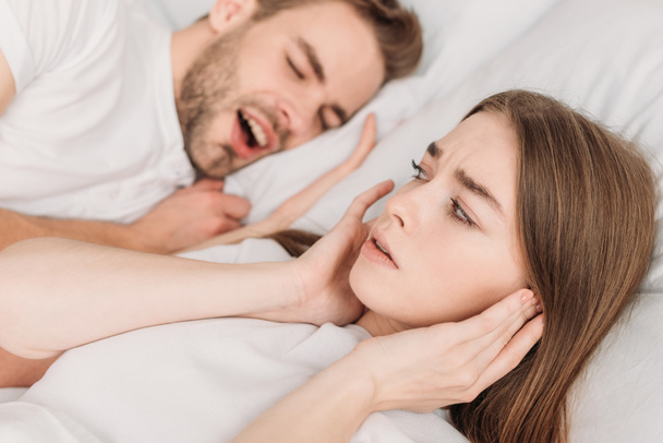 いびきの夫の近くのベッドに横たわっている間に手で耳を差し込む女性の選択的な焦点 - 写真・画像