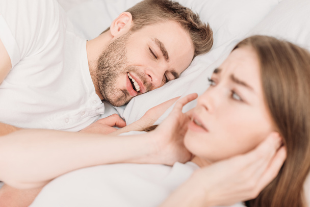 selective focus of exhausted woman plugging ears with hands while lying in bed near snoring husband - Fotografie, Obrázek