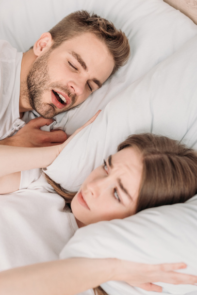 awakened woman plugging ears with pillow while lying in bed near snoring husband - 写真・画像