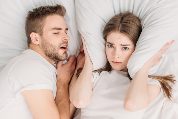top view of woman plugging ears with pillow while lying in bed near snoring husband - Fotó, kép