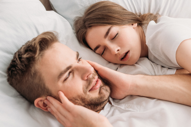 exhausted man plugging ears with hands while lying in bed near snoring wife - Photo, Image
