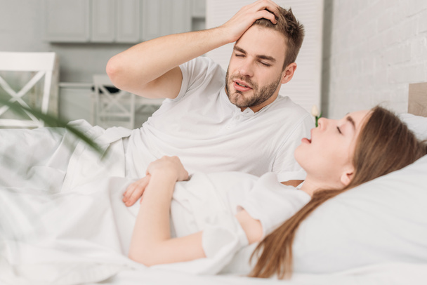 Erschöpfter Mann hält Hand auf Kopf, während er neben schnarchender Frau im Bett liegt - Foto, Bild