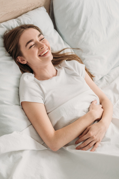 cheerful girl laughing while lying in bed and holding hands on stomach - Zdjęcie, obraz