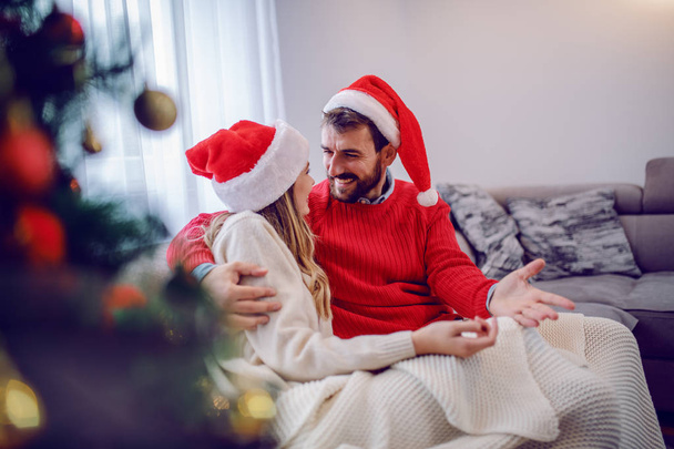 Charmant couple caucasien en chandails et avec chapeau de Père Noël sur les têtes assis sur le canapé dans le salon à côté de l'arbre de Noël et profiter de leur temps
. - Photo, image
