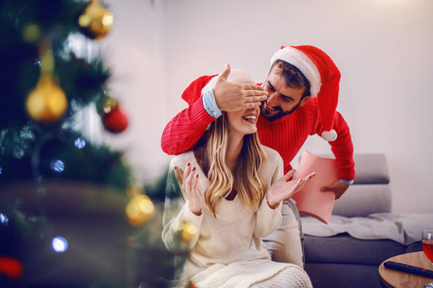 Handsome caucasian man in sweater and with santa hat on head covering his girlfriend's eyes and hiding present behind back. Christmas holidays. - Photo, Image