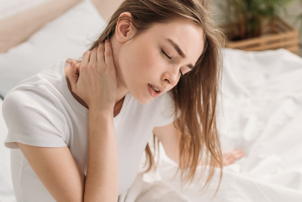 young woman sitting in bed with closed eyes and suffering from neck pain - Foto, afbeelding