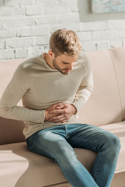 young man sitting on sofa at home and suffering from stomach pain - Φωτογραφία, εικόνα