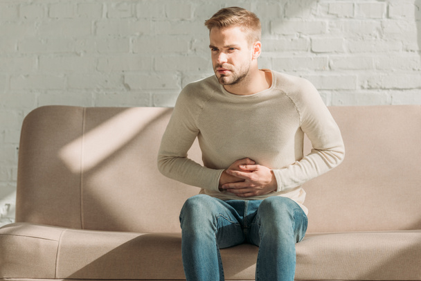 upset man sitting on sofa and touching stomach while suffering from abdominal pain - Φωτογραφία, εικόνα