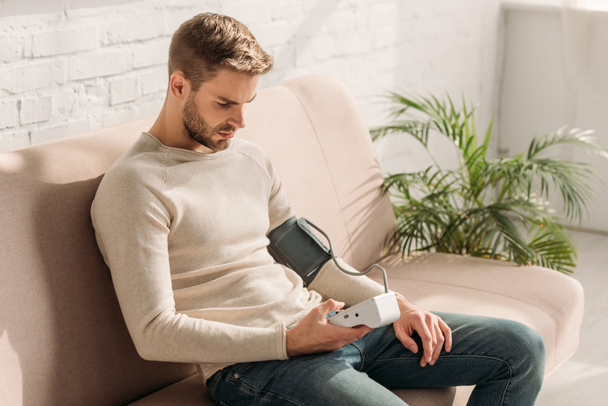 handsome man sitting on sofa at home and measuring blood pressure with tonometer - Photo, Image