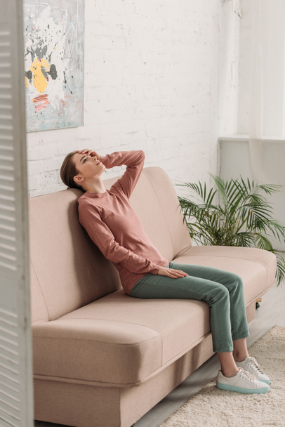 Nuori nainen istuu sohvalla ja etsii ylös kärsiessään päänsärky
 - Valokuva, kuva