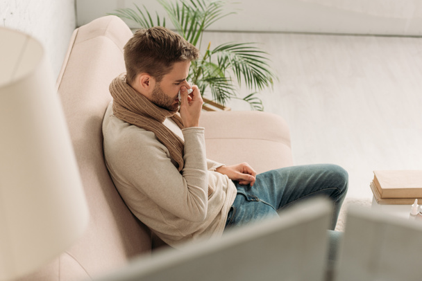 selective focus of sick man sitting on sofa and sneezing in napkin - Φωτογραφία, εικόνα