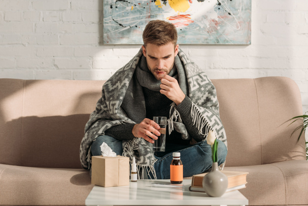 ill man, wrapped in blanket, sitting on sofa and taking medicines - Foto, Imagem