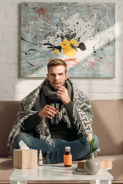 sick man, wrapped in blanket, sitting on sofa and taking medicines - Foto, Imagem