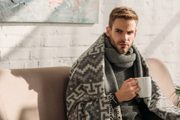 sick, unhappy man, wrapped in blanket, looking away while holding cup of warming drink - Photo, Image