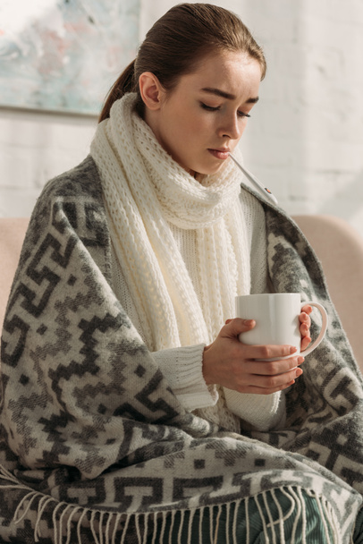 sick woman with warm scarf around neck, wrapped in blanket, holding cup of tea - Fotoğraf, Görsel