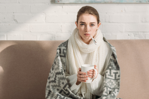 sick girl looking at camera while measuring temperature and holding cup of warming drink - Photo, Image