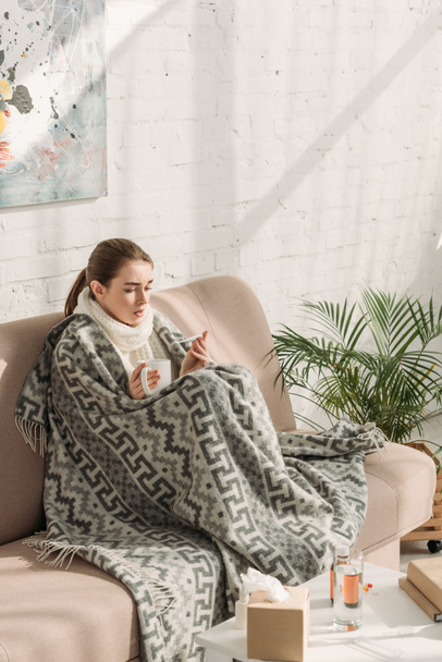 sick girl, wrapped in blanket, looking at thermometer and holding cup of warming drink - Foto, Bild