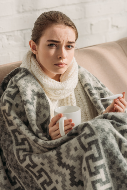 Beteg lány, takaróba csomagolva, kamerába néz, miközben hőmérőt és egy csésze melegítő italt tart a kezében. - Fotó, kép