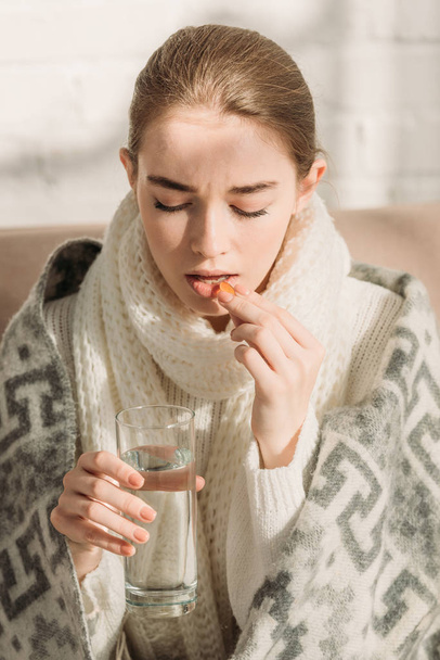 sick girl, wrapped in blanket, holding glass of water while taking medicine - Photo, Image