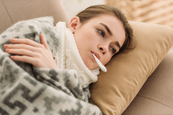 mulher doente deitado no sofá sob cobertor e medir a temperatura
 - Foto, Imagem