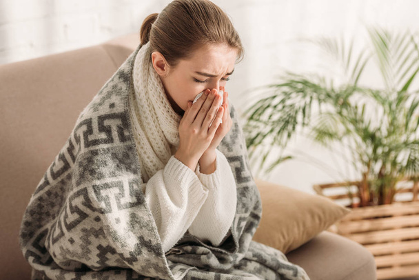 sick woman sneezing in napkin while sitting on sofa and wrapping in blanket - Zdjęcie, obraz
