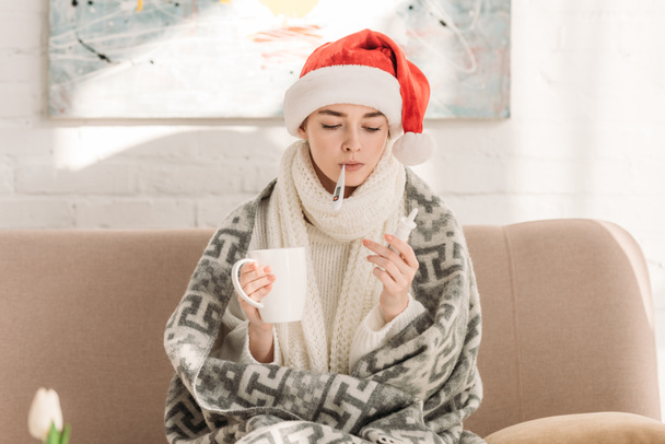 sick girl in santa hat measuring temperature while holding nasal spray and cup of warming drink - Photo, Image
