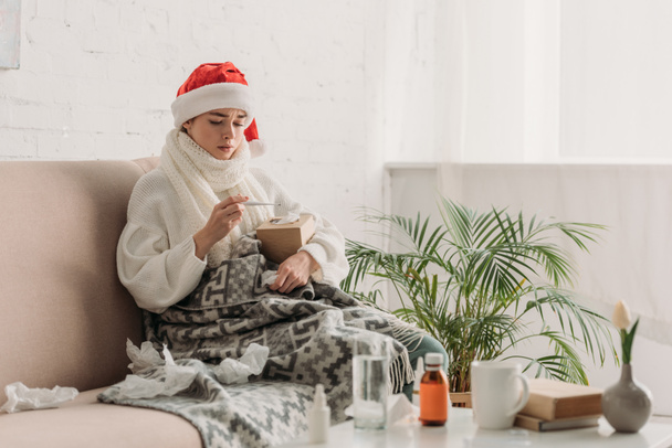 sick woman in santa hat, wrapped in blanket, sitting on sofa near table with medicines and looking at thermometer - Photo, Image