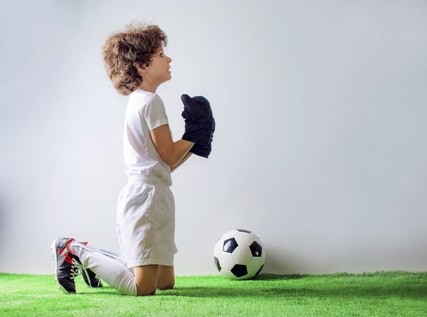 Cute cheerful curly-haired boy soccer goalkeeper on his knees, hands clasped in prayer and looking into the empty space. Gray background. - Fotó, kép