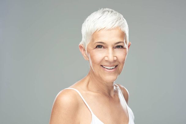 schöne lächelnde Seniorin mit kurzen grauen Haaren, die vor grauem Hintergrund posiert. Schönheitsfotografie. - Foto, Bild