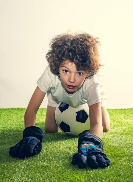 A boy crawling into the camera. Football classes for children. Portrait of cute shaggy boy with soccer ball lying over grassy studio background, free space - Fotó, kép