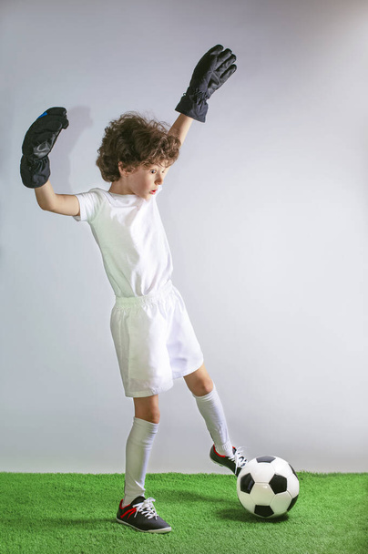 Kids - soccer champion. Boy goalkeeper with a surprised face in football sportswear with ball on gray background. Sport concept.Copy space. - 写真・画像