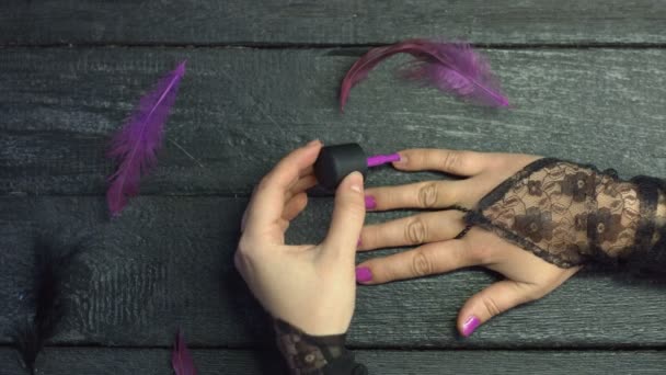 Manicure.Girl ζωγραφίζει τα νύχια της με μωβ βερνίκι τζελ σε μαύρο ξύλινο φόντο ανάμεσα σε μωβ φτερά. Χαριτωμένα χέρια με ένα όμορφο μανικιούρ στα νύχια.  - Πλάνα, βίντεο