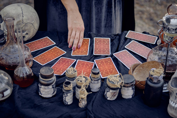 Hexe in schwarzem Gewand, die auf Karten, die am Tisch stehen, mit Zubehör für Zauber, Magie und Hexerei zaubert, während sie im Kiefernwald posiert. Nahaufnahme. - Foto, Bild
