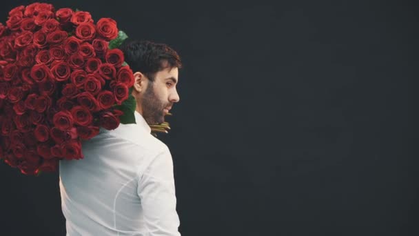 Handsome man with large bouquet of roses on the shoulder standing his back to the camera, turning around, smiling, raising eyebrow, looking seducing. - Footage, Video
