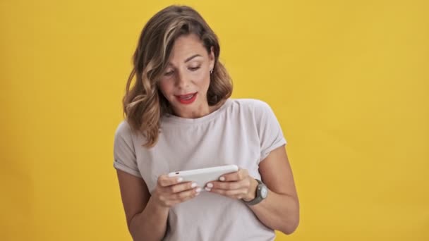 Pretty woman with red lips playing on her smatphone while holding it horizontally and smiles over yellow background isolated - Video
