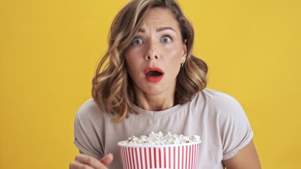 Scared and frighten young woman with red lips holding a basket of popcorn while looking at the camera over yellow background isolated - Záběry, video