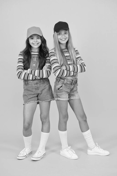 Fashion inspired by the sneaker culture. Happy kids keeping arms crossed with fashion look. Fashion small girls in casual wear smiling on yellow background. Streetwear fashion for little children - 写真・画像