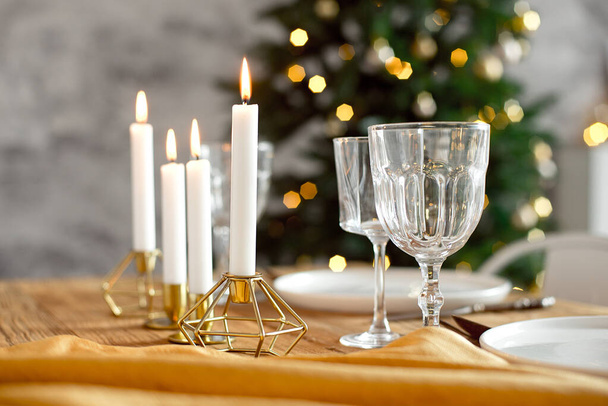 Table is served for a Christmas dinner in the living room, close-up view, table setting with a golden tablecloth, white plates, forks, knives, glasses for wine, candles in gold candlesticks, Christmas - Foto, Imagen