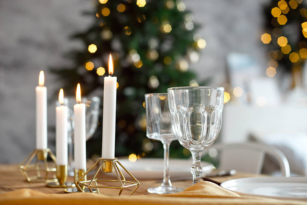 Table is served for a Christmas dinner in the living room, close-up view, table setting with a golden tablecloth, white plates, forks, knives, glasses for wine, candles in gold candlesticks, Christmas - Фото, изображение