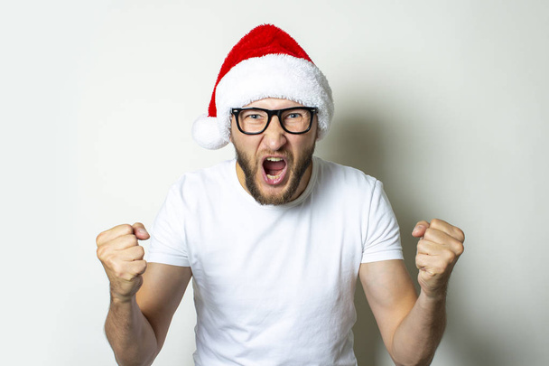 Young man in glasses and a Santa Claus hat shouts on a white background. Christmas concept. Gesture victory, super, cheers, joy - 写真・画像