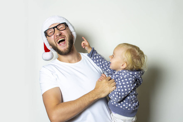Un joven con gafas y un sombrero de Papá Noel juega con un niño sobre un fondo blanco. Concepto de Navidad, celebración en familia, vacaciones en familia
 - Foto, Imagen