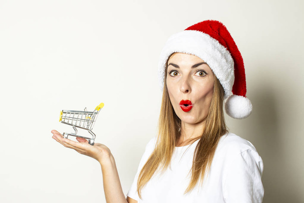 Uma jovem mulher com um rosto surpreso em um chapéu de Papai Noel está segurando um carrinho de compras em suas mãos em um fundo branco. Conceito de venda de Natal, sexta-feira preta, desconto
 - Foto, Imagem