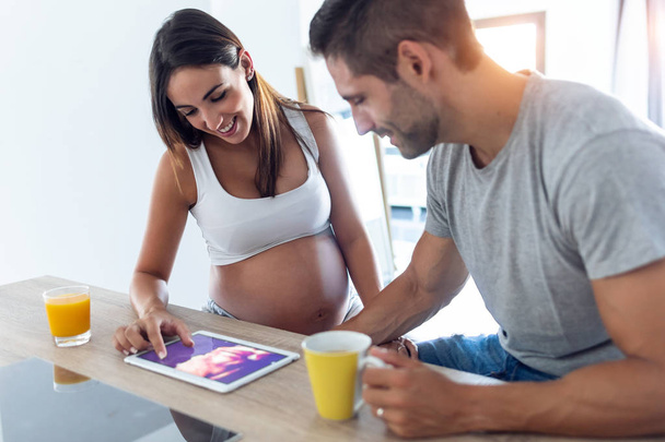 Pěkný mladý těhotný pár při pohledu na ultrazvuk jejich dítěte na digitální tablet v kuchyni doma. - Fotografie, Obrázek