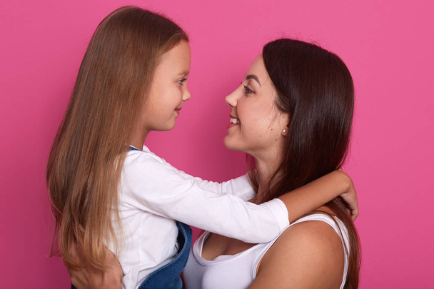 Excited mother and cute kid girl looking on each other with charming smiles, weaeing white shirts, having long hair, posing isolated over pink background, adorable female misses her liitle daughter. - Fotoğraf, Görsel