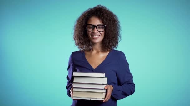 African student with glasses on blue background in the studio holds stack of university books from library. Girl smiles, she is happy to graduate. - Video