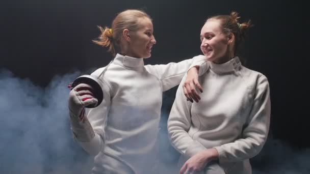 Две молодые женщины-фехтовальщицы в белых защитных костюмах разговаривают друг с другом - опираясь на плечо друга и держа в руках меч
 - Кадры, видео
