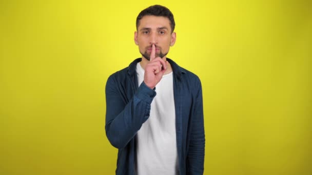 Een jongeman in een blauw shirt en wit T-shirt kijkt naar de camera en houdt een vinger aan haar lippen die een teken van stilte en geheim toont. Gele achtergrond met kopieerruimte. 4k slow motion beelden - Video