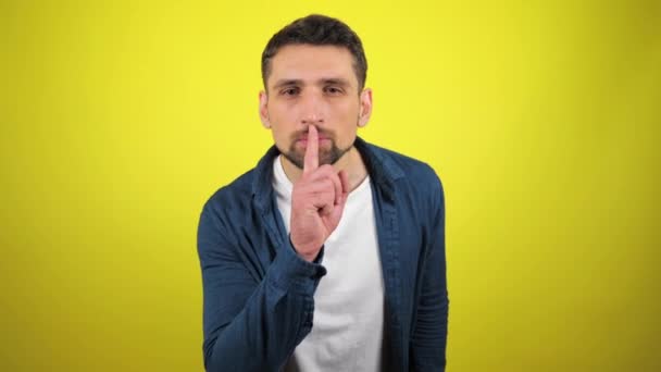 Een jongeman in een blauw shirt en wit T-shirt kijkt naar de camera en houdt een vinger aan haar lippen die een teken van stilte en geheim toont. Gele achtergrond met kopieerruimte. 4k slow motion beelden - Video