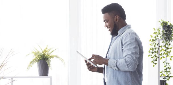 Usmívající se africký muž pomocí digitálního tabletu, zatímco stojí u okna - Fotografie, Obrázek